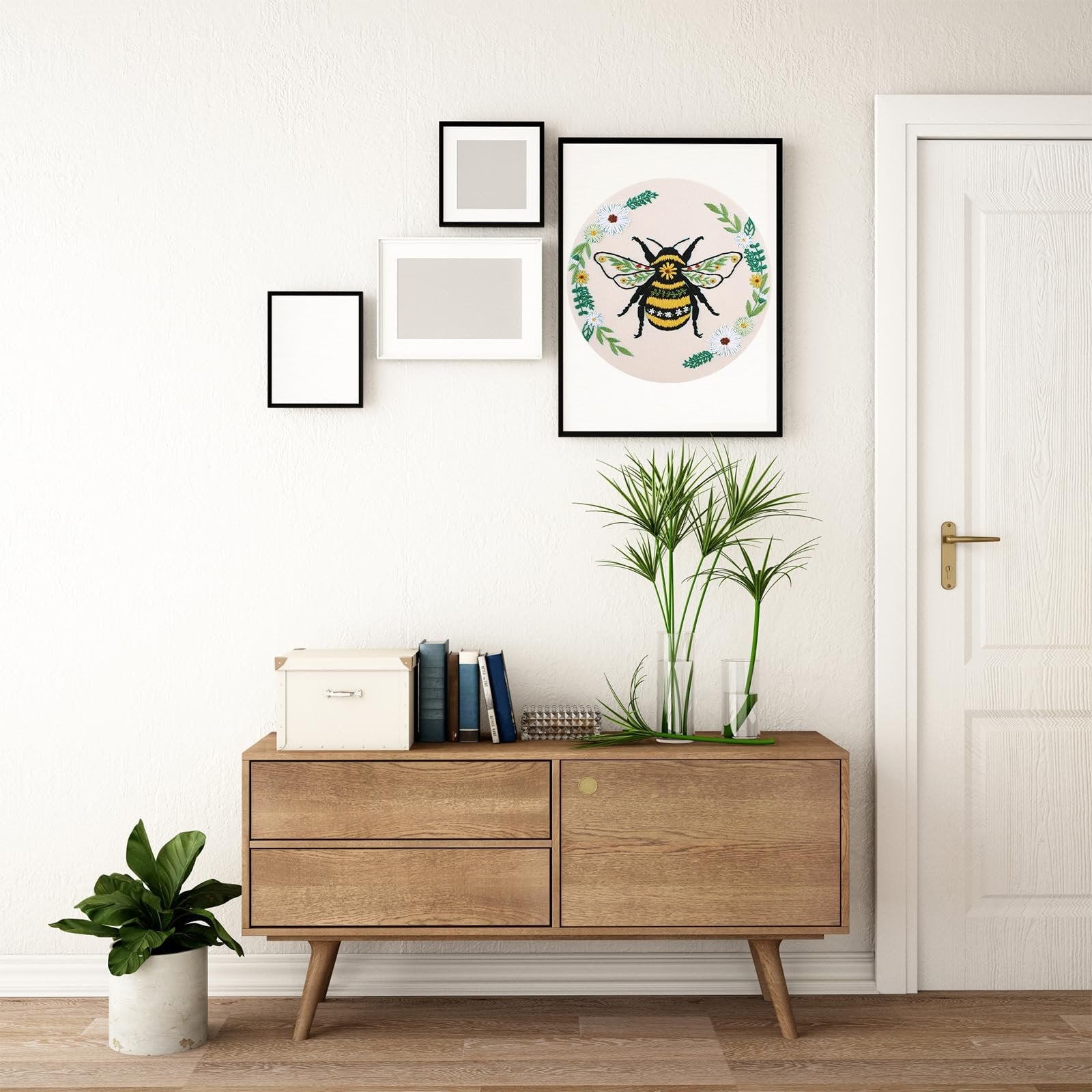 Embroidery art of a bee with flowers in a framed picture on a wall above a wooden cabinet.
