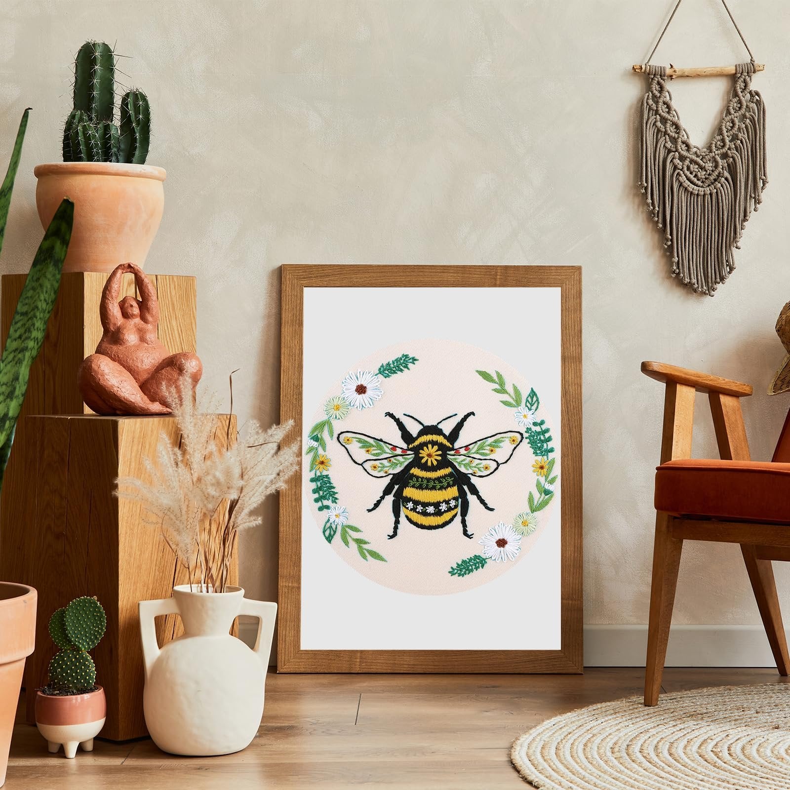 Embroidery hoop with a bee and floral wreath design, displayed in a wooden frame on a shelf with plants and decor.