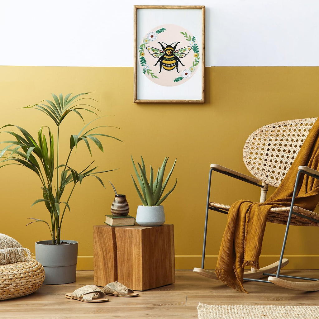 Embroidery hoop art with a bee and flowers hanging on a yellow and white wall above plants and a chair