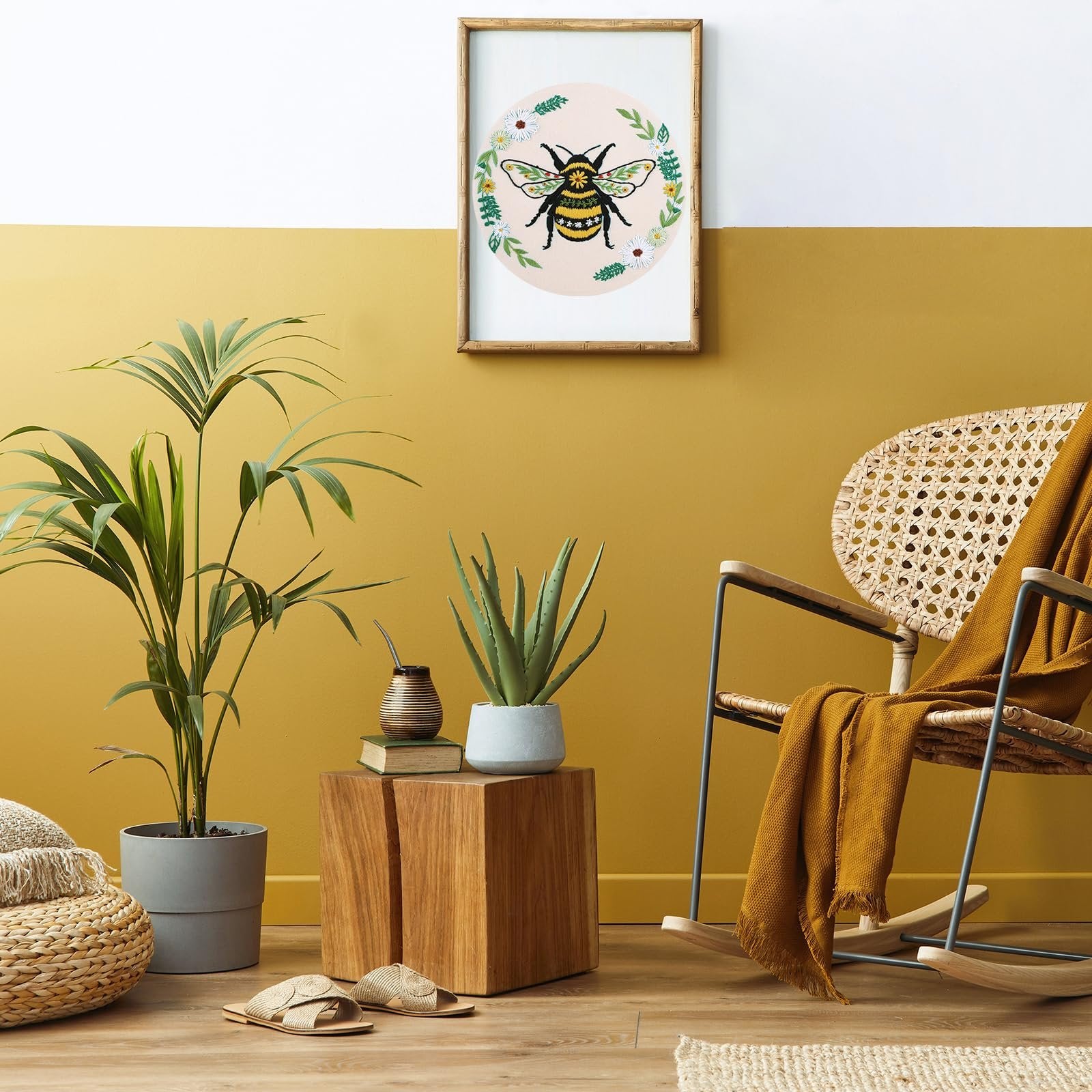 Embroidery hoop art with a bee and flowers hanging on a yellow and white wall above plants and a chair