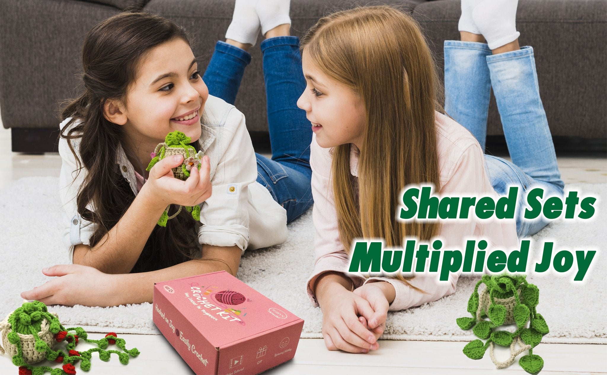 Two young girls lying on a rug, smiling and holding crocheted succulent plant decorations. A pink crochet kit box is in the foreground.