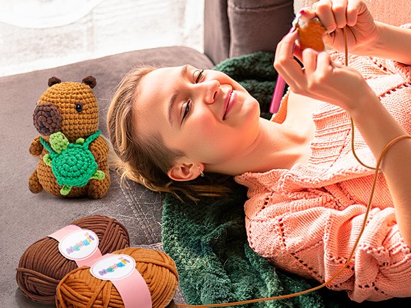 Young woman crocheting a capybara plush toy with a turtle on its back, yarn balls nearby