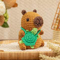 Crocheted capybara toy holding a green turtle, displayed on a wooden tray with decorative elements.