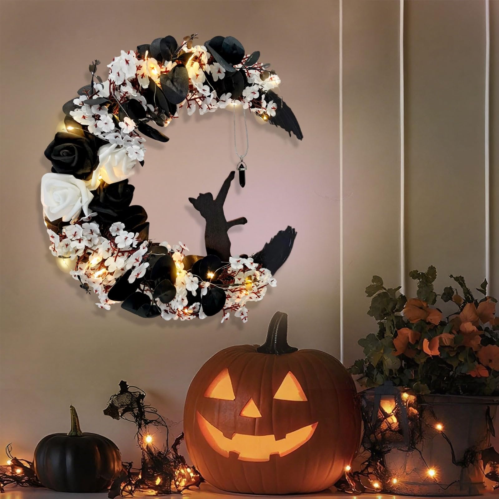 Black cat crescent moon Halloween wreath with lights, black and white roses, and a hanging pendant, displayed with pumpkins and fall foliage.
