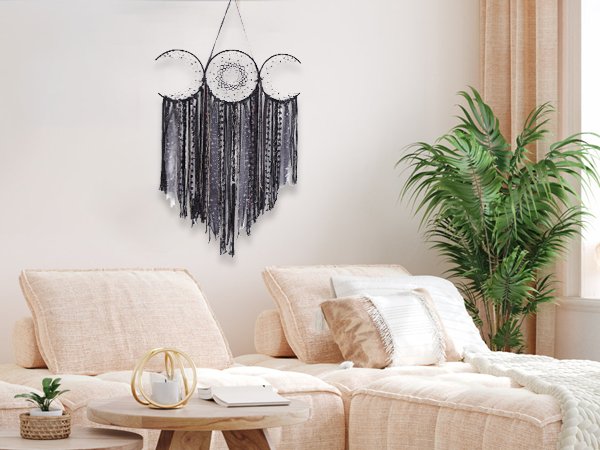 Black triple moon macrame wall hanging with mixed textile fringe displayed on a wall above a sofa.