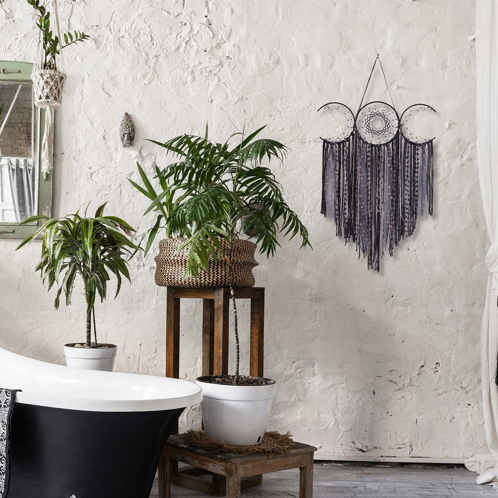 Black triple moon macrame wall hanging with crescent moons and textile fringe, displayed in a bathroom setting with plants and a bathtub.