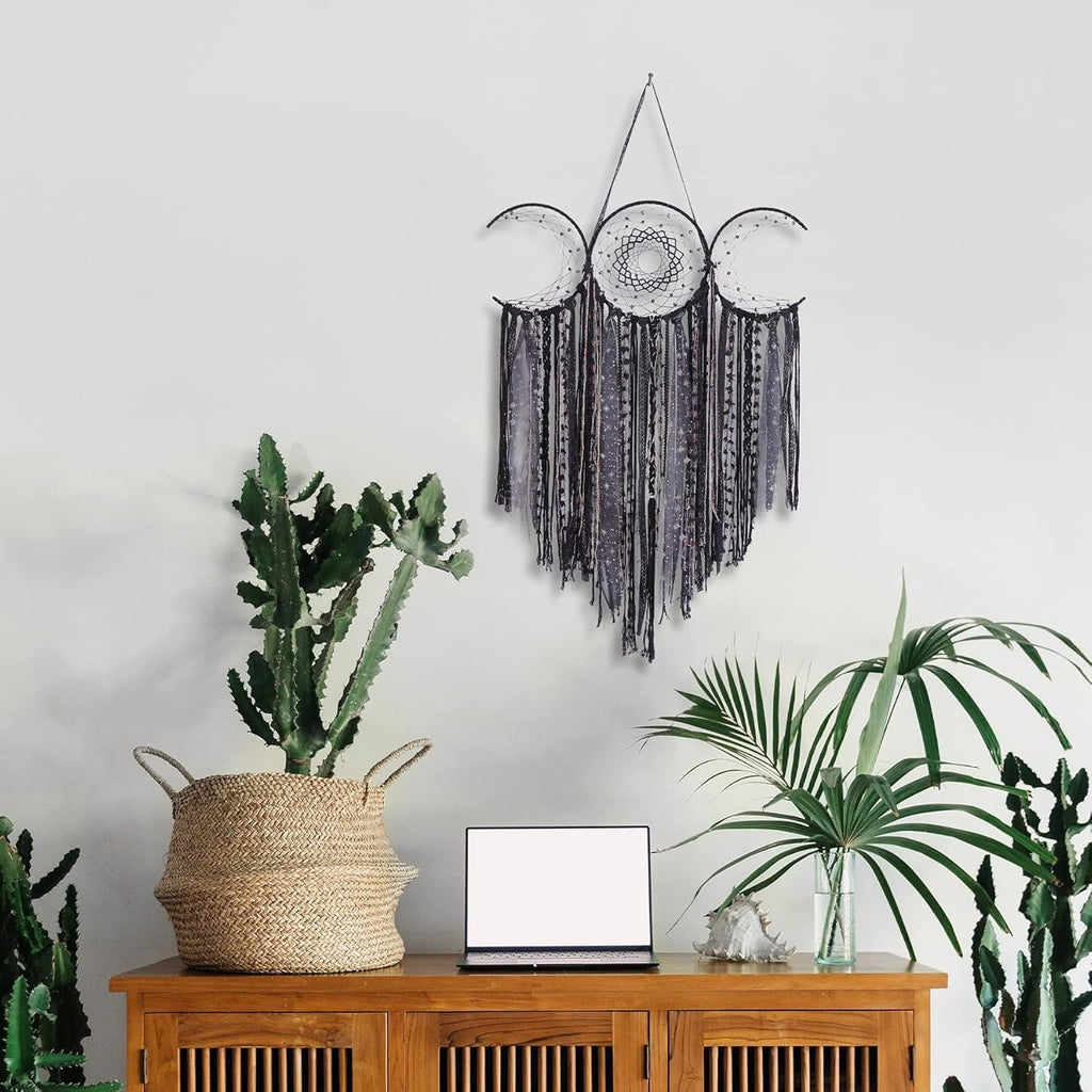 Black triple moon macrame wall hanging with crescent dream catchers and textile fringe, displayed above a wooden cabinet with plants and a laptop.