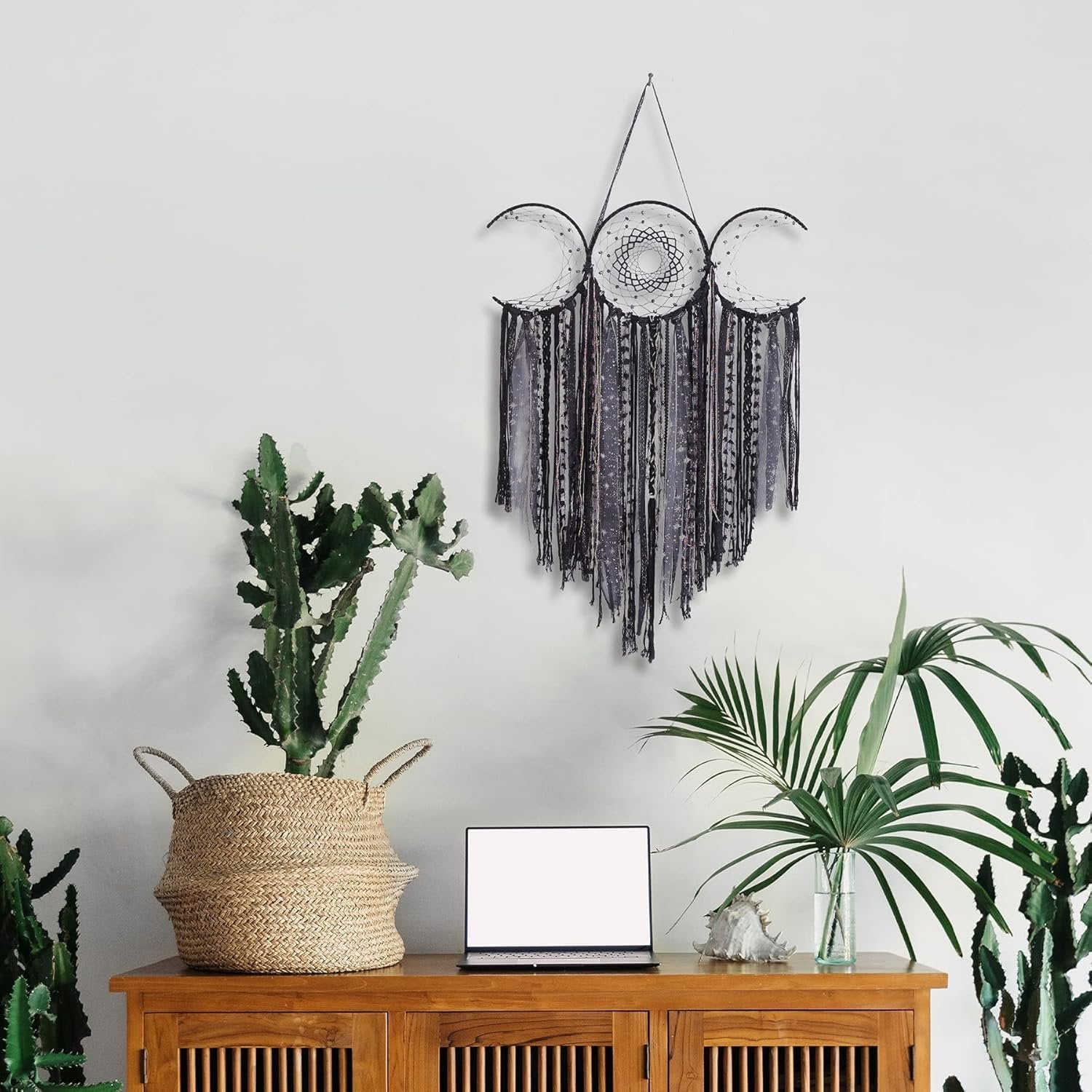 Black triple moon macrame wall hanging with crescent dream catchers and textile fringe, displayed above a wooden cabinet with plants and a laptop.