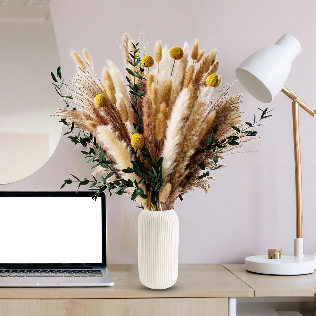 Boho dried flower bouquet with pampas grass, eucalyptus, and craspedia in a ribbed vase on a desk with a laptop and lamp.