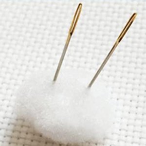 Two sewing needles stuck in a white cotton ball on pre-printed cross-stitch fabric.