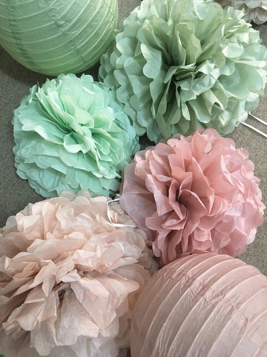 Sage green and rose pink tissue paper pom poms and paper lanterns arranged on a textured surface.