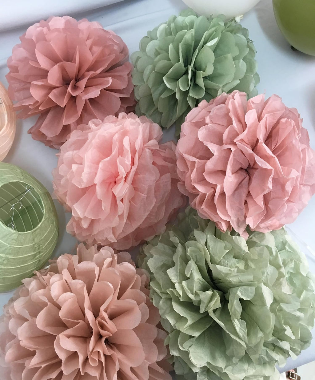 Sage green and rose pink tissue paper pom poms and paper lanterns arranged on a white surface.