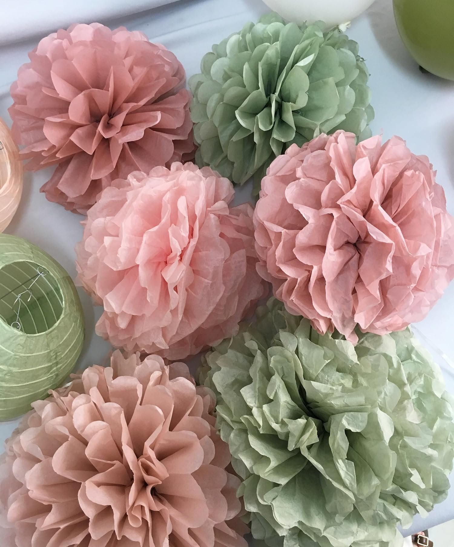 Sage green and rose pink tissue paper pom poms and paper lanterns arranged on a white surface.