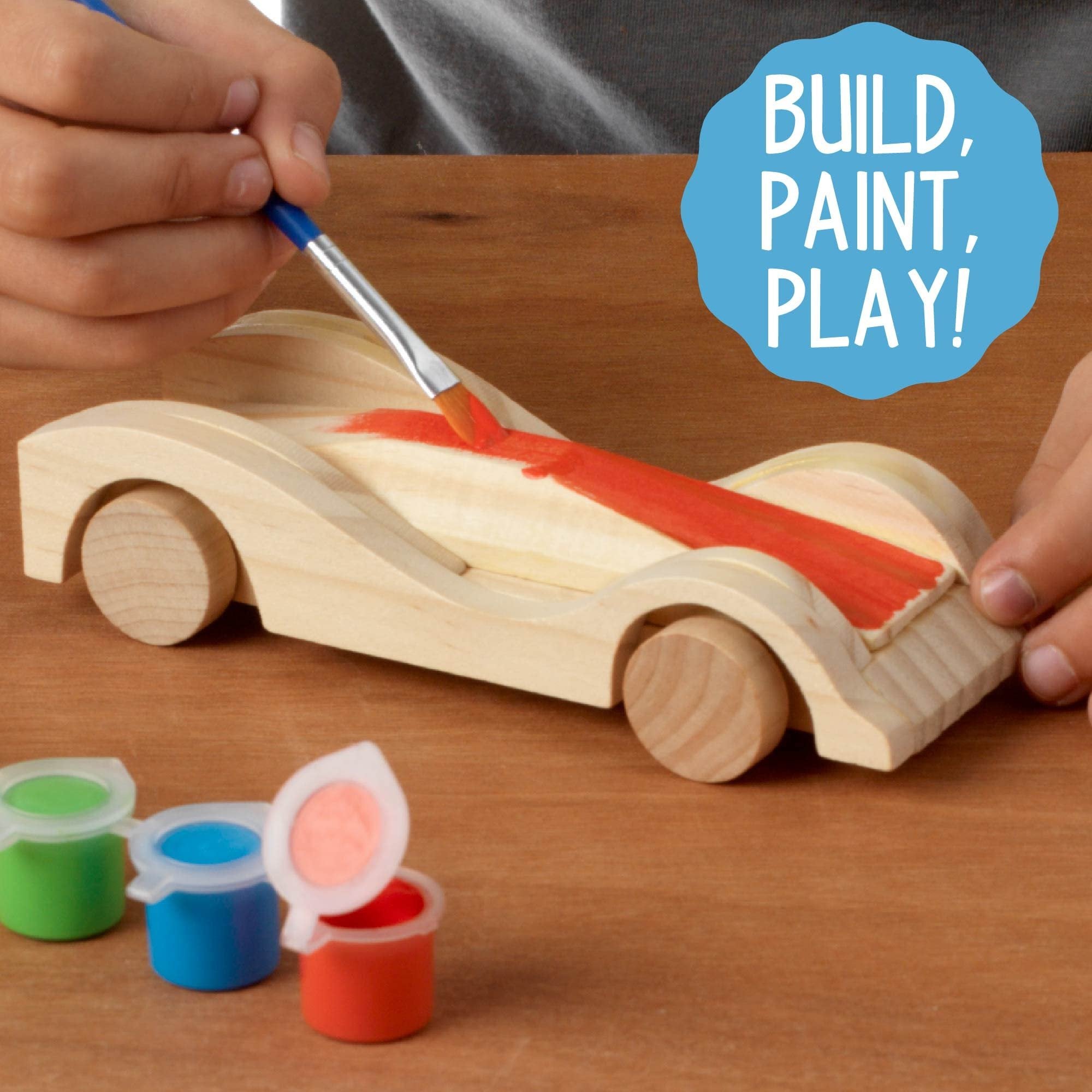 Child's hands painting a wooden race car with red paint, small paint pots nearby.