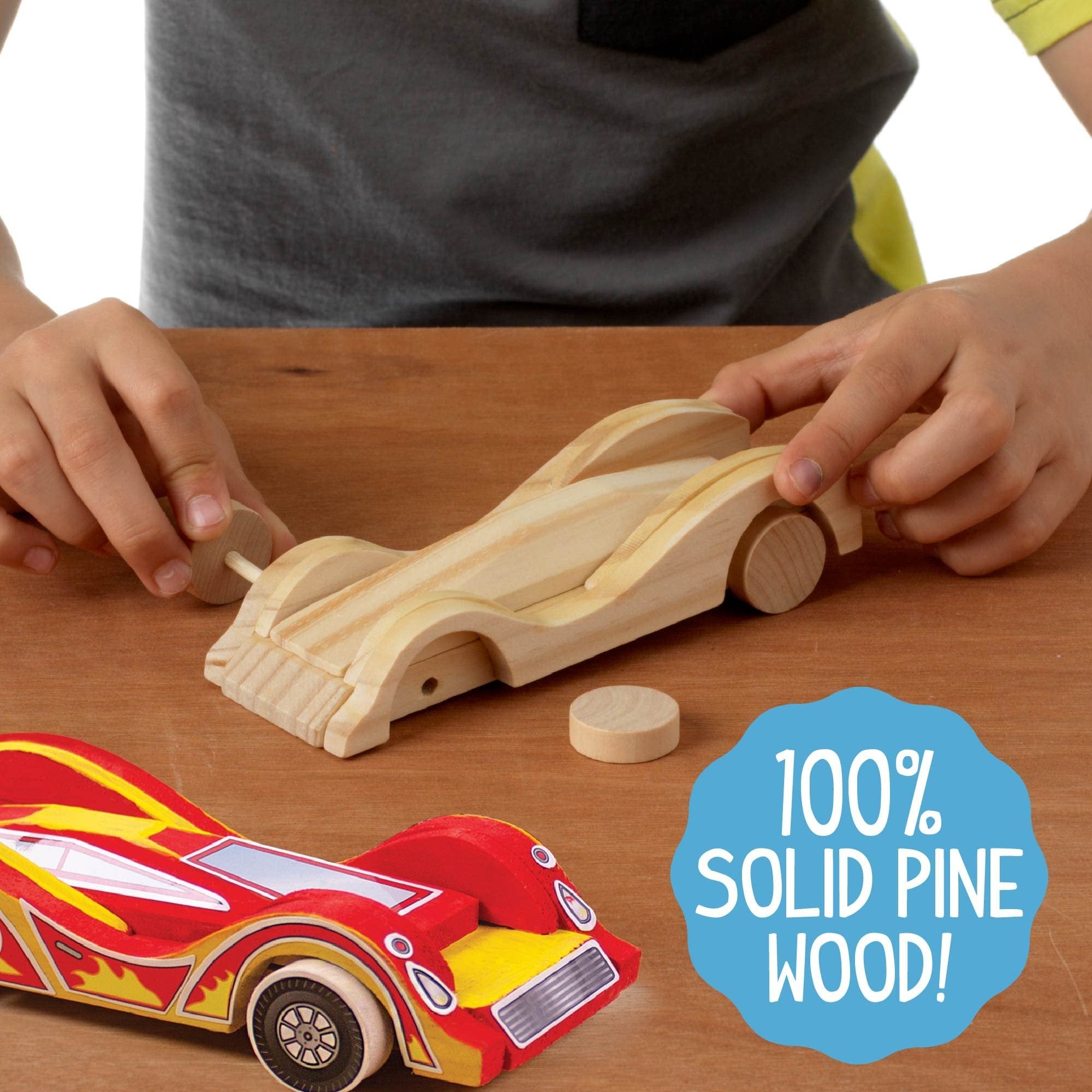Child's hands assembling a wooden race car kit on a wooden surface, with a painted car in the foreground.