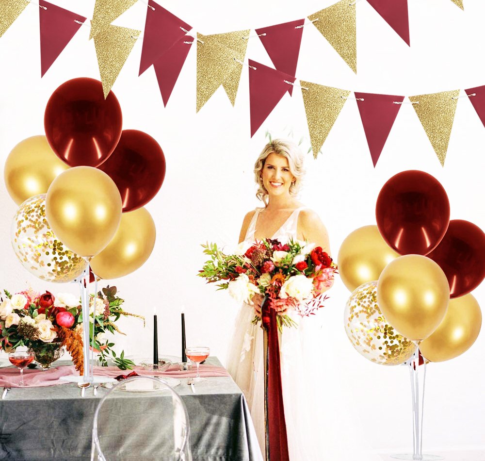 Woman in white dress holding flowers with burgundy and gold balloons and bunting decor