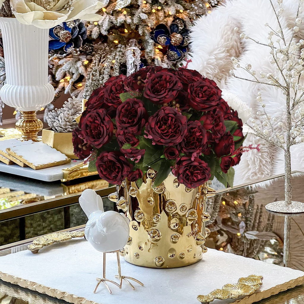 Burgundy artificial peony bouquet in a gold textured vase, with a white bird figurine and festive decor in the background.