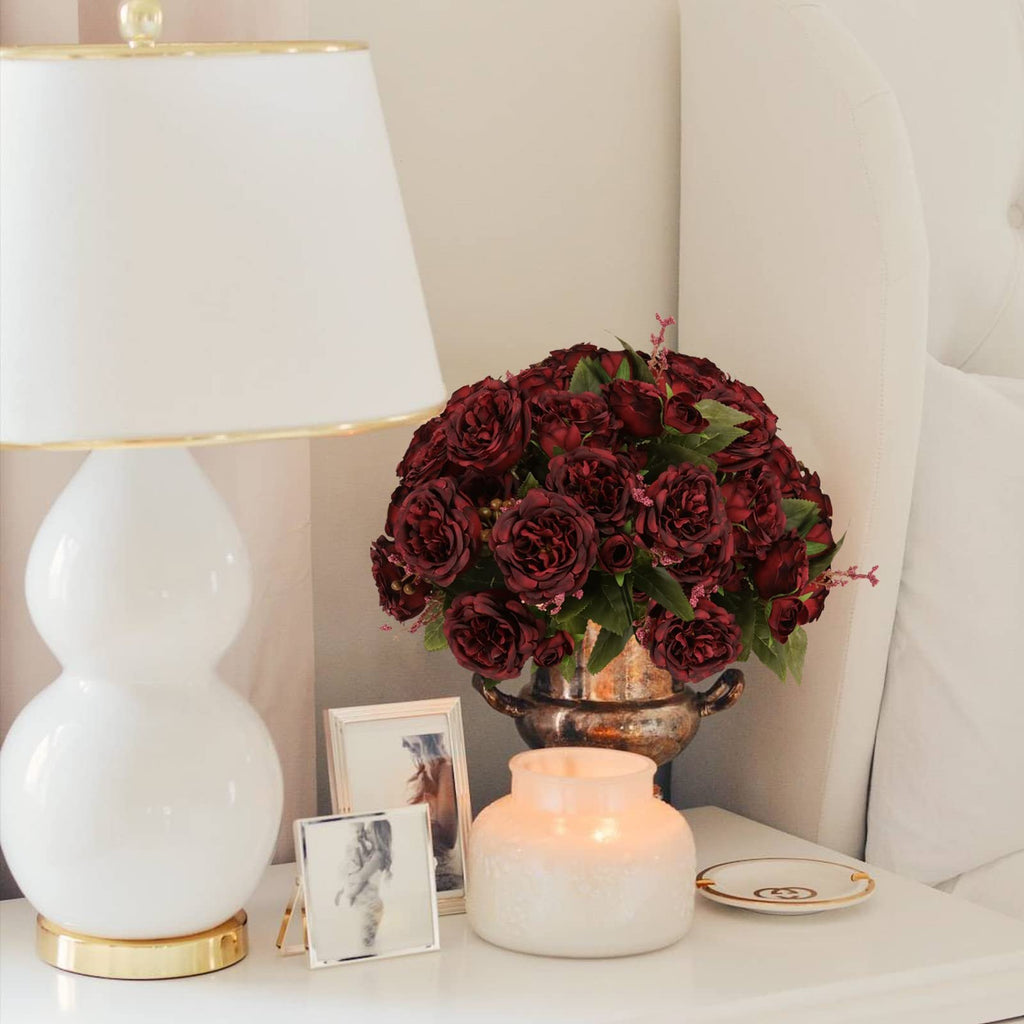 Burgundy artificial peony bouquet in a decorative vase on a nightstand next to a lamp and framed photos.