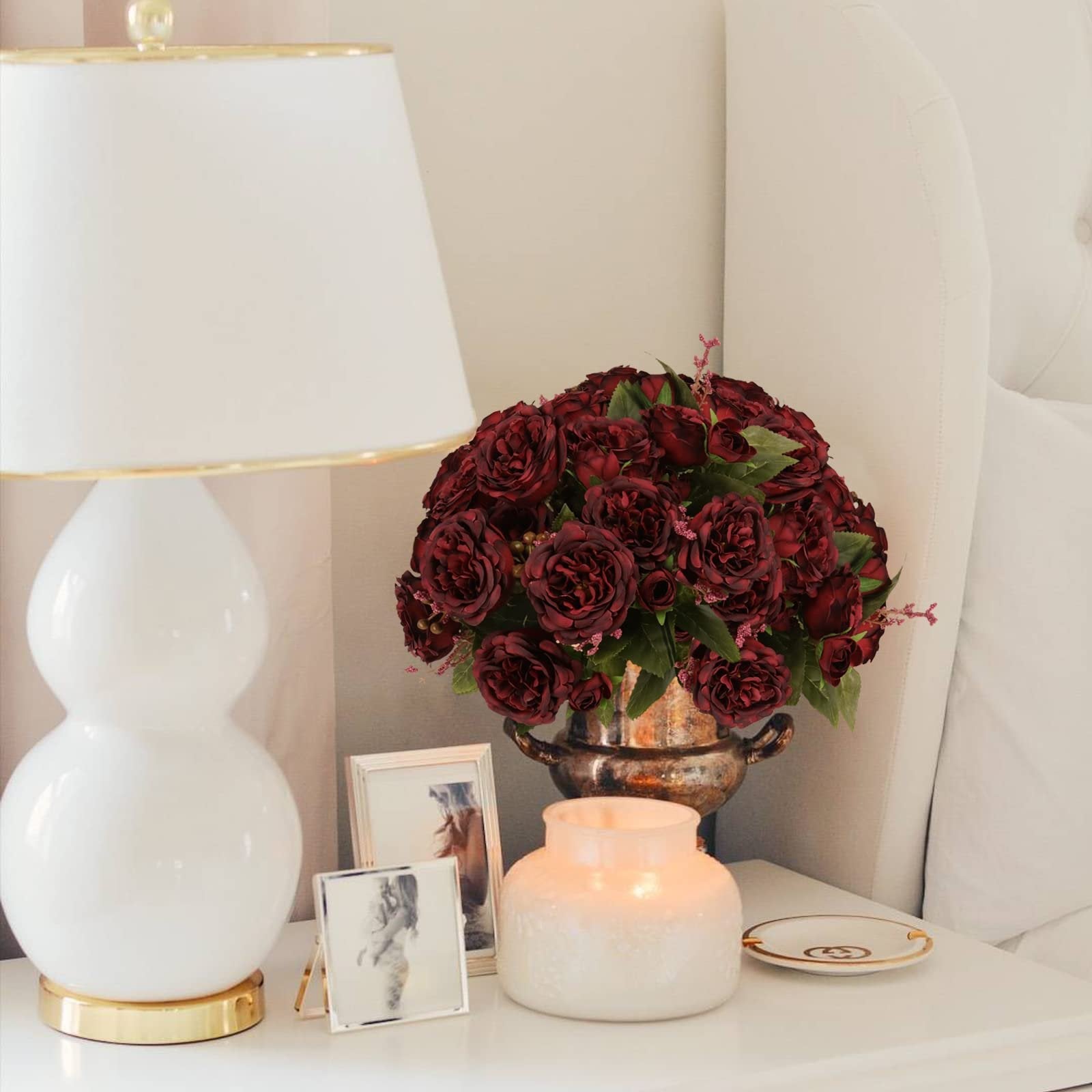 Burgundy artificial peony bouquet in a decorative vase on a nightstand next to a lamp and framed photos.