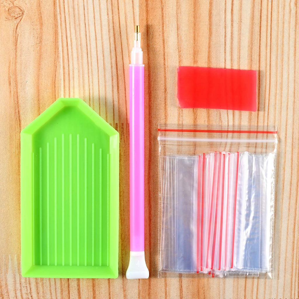 Diamond painting kit accessories including a green tray, pink drill pen, red wax, and multiple small plastic bags on a wooden background.