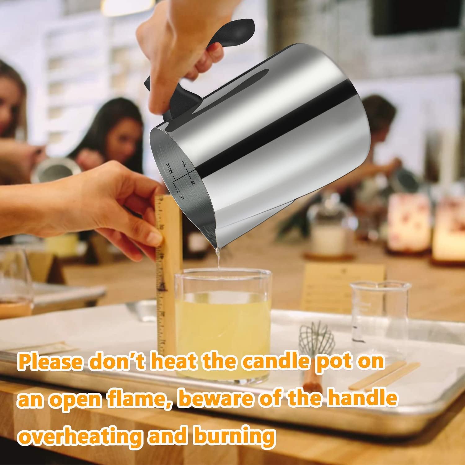 Hand pouring liquid from a stainless steel pitcher into a glass, with a ruler for scale. Candle making supplies on a tray.
