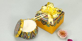 Candle in a decorative tin with lid open, next to a gift box with a yellow ribbon.