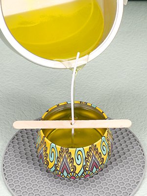 Pouring melted yellow wax into a decorative yellow tin with a wick centered by a wooden stick.