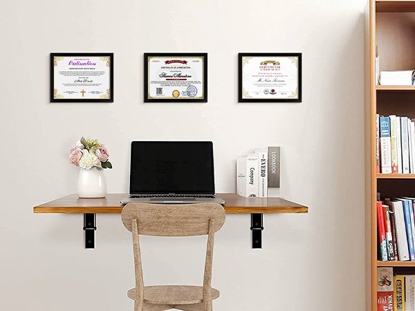 Three framed certificates displayed on a wall above a wooden desk with a laptop and vase of flowers.