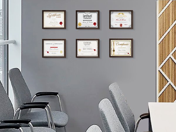 Six framed certificates displayed on a gray wall above chairs in an office setting.