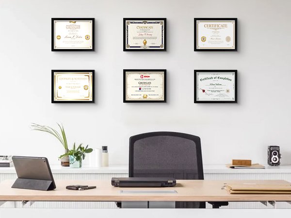 Six framed certificates displayed on a wall above a desk with a laptop and tablet.