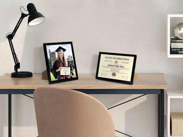 Two black certificate frames on a wooden desk with a desk lamp and a chair. One frame shows a graduate holding a diploma.