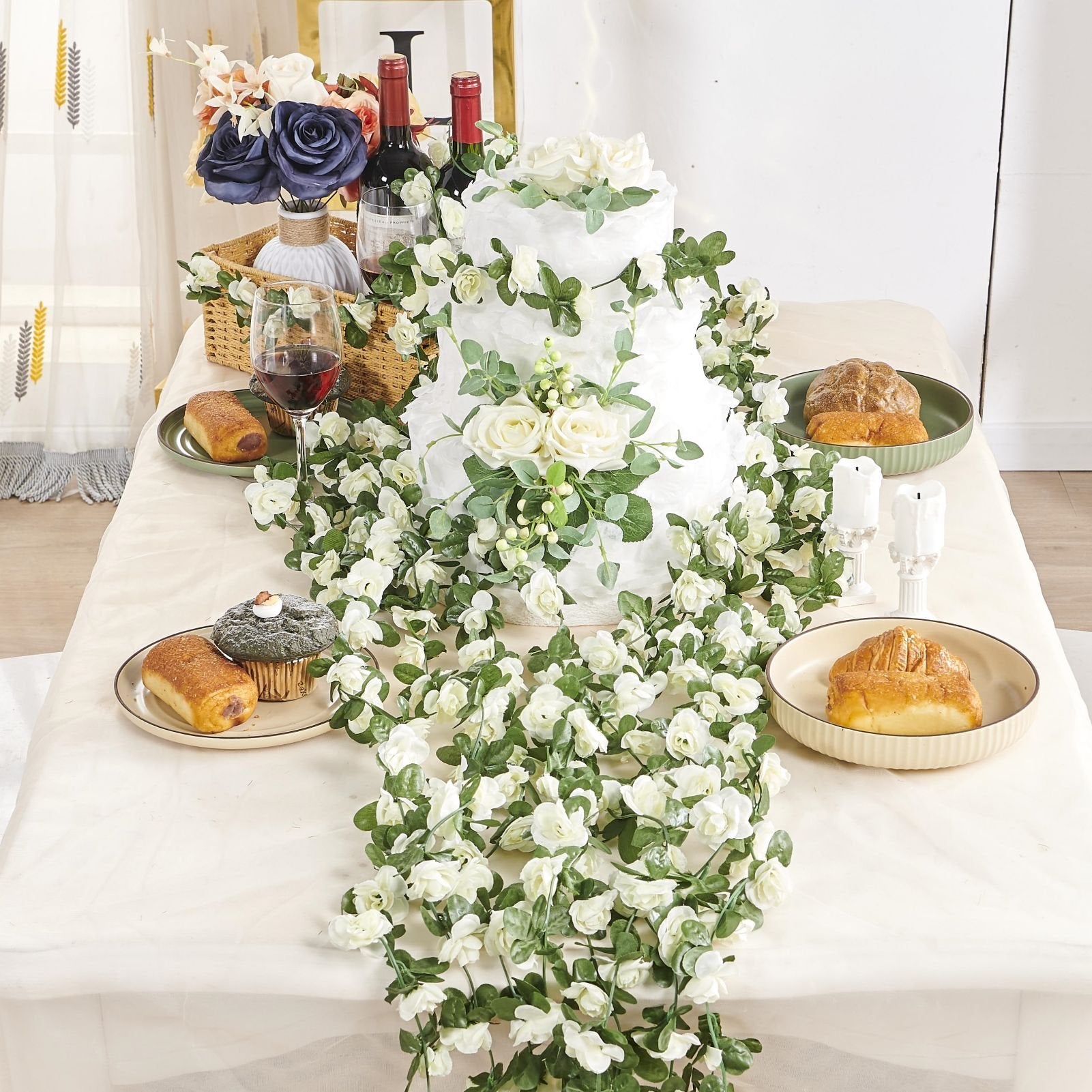 Champagne artificial rose vine garland draped over a tiered cake and table setting with bread and wine.