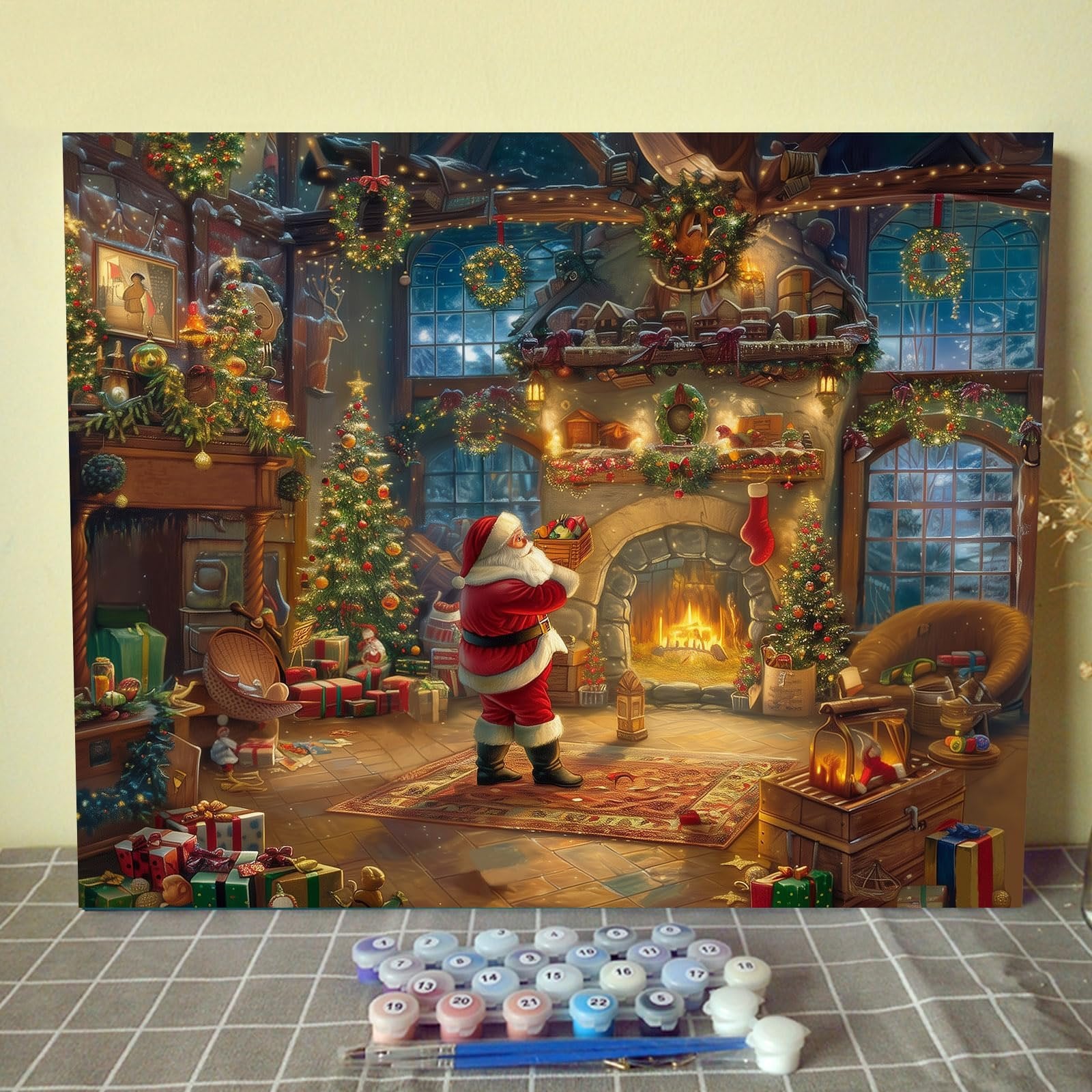 Christmas scene painting with Santa by a fireplace, surrounded by trees and gifts, with paint pots and brushes in the foreground.