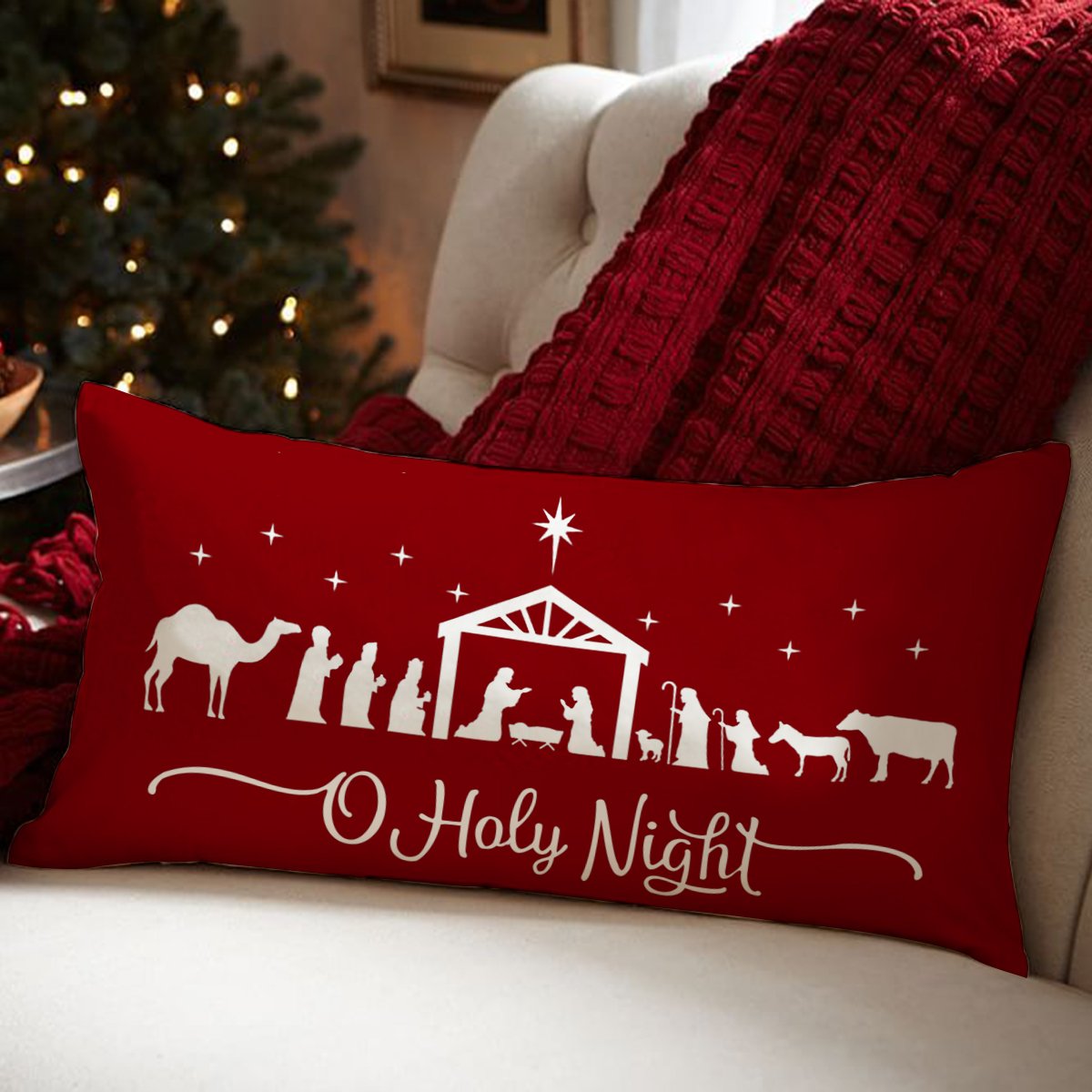 Red Christmas pillow cover with white nativity scene and "O Holy Night" text, resting on a sofa with a red blanket and Christmas tree in background.