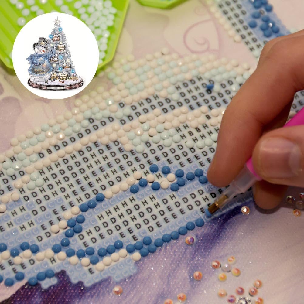 Diamond painting kit in progress showing a hand using a pink tool to place rhinestones on a numbered canvas with a small inset image of the finished Christmas tree and snowman design.