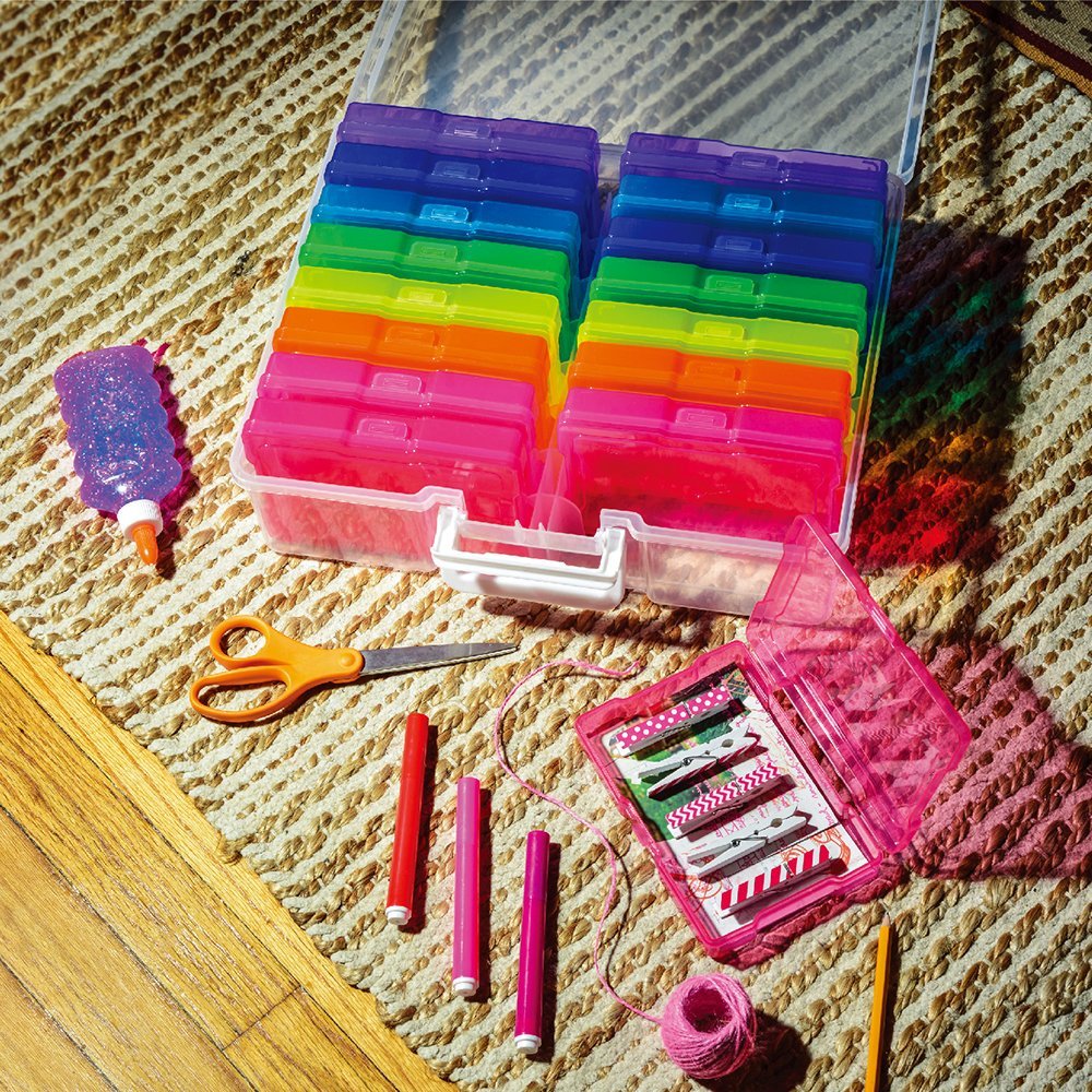 Open clear photo storage box with rainbow colored cases, scissors, glitter glue, markers, and clothespins on a rug.