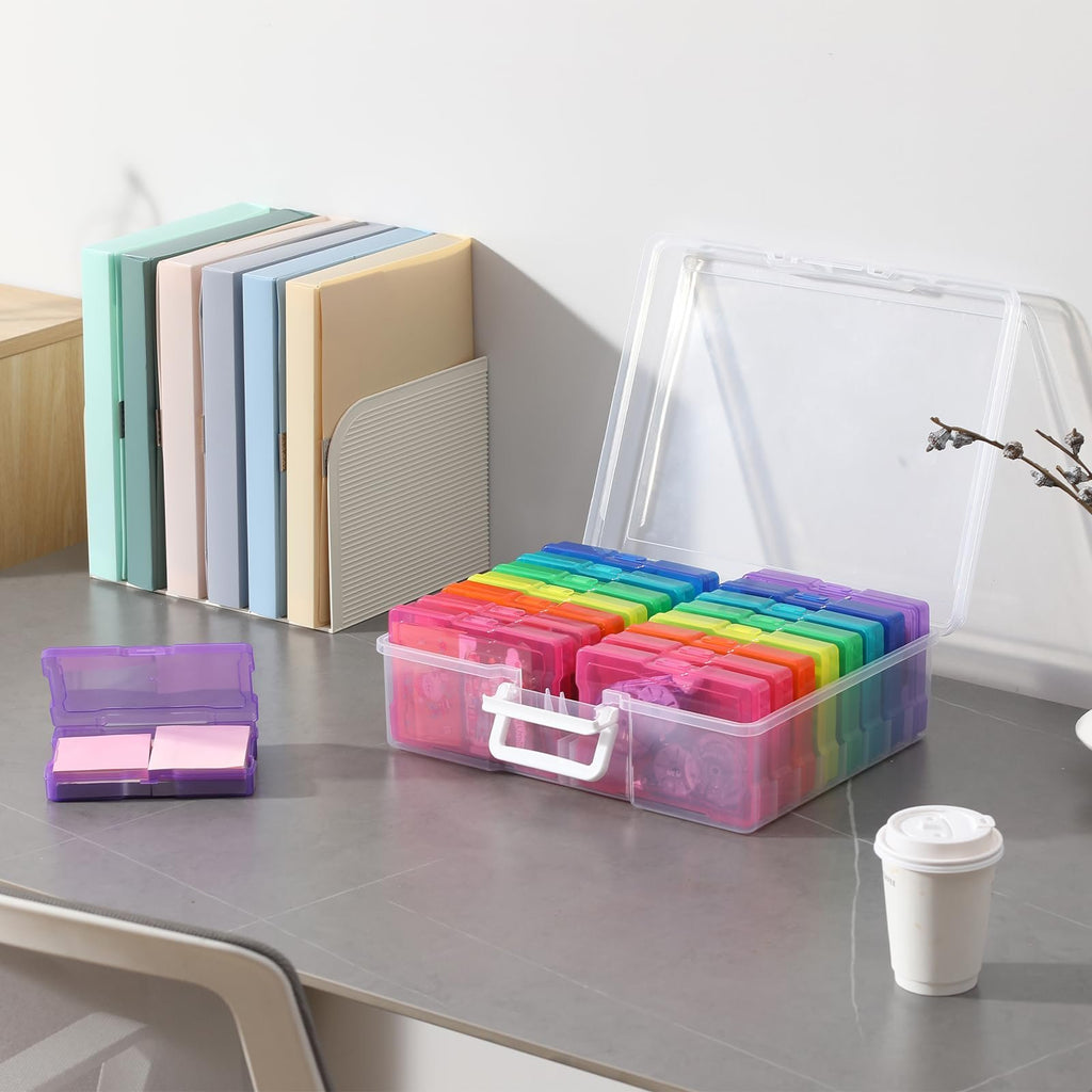 Clear photo storage box with handle, open, showing 16 rainbow-colored cases inside, on a desk with files and a small purple case.