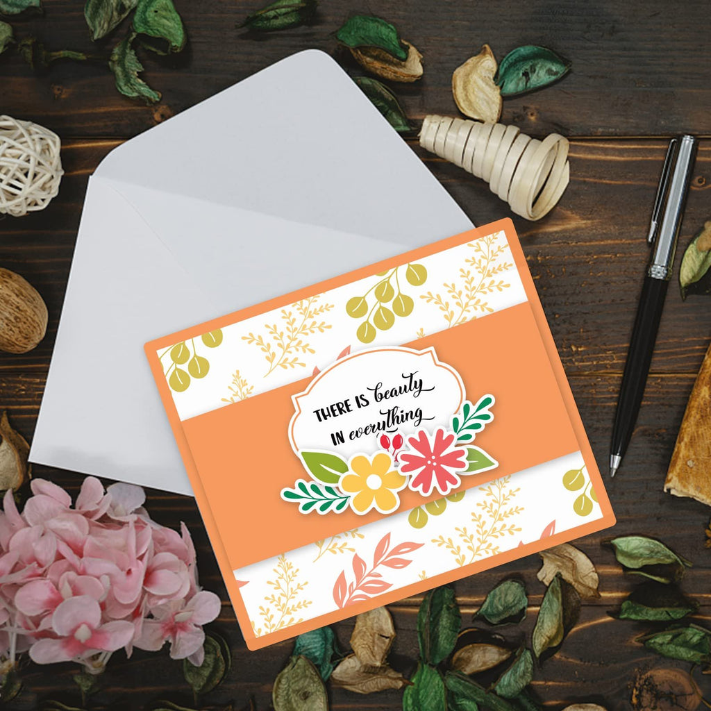 Card making supplies with envelope, pen, and decorative elements on a wooden surface. Features a card with floral design and text.