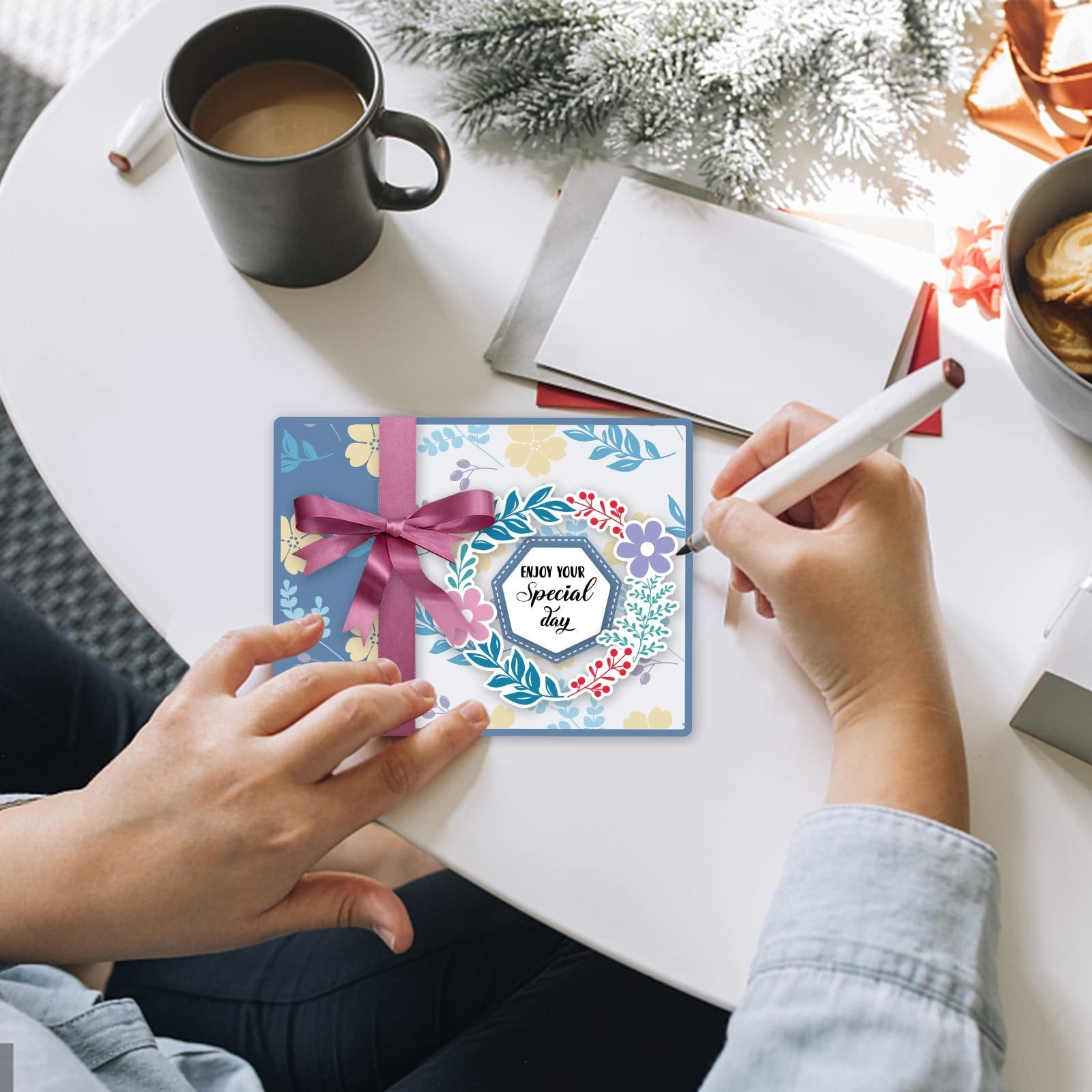 Person writing on a card with floral design and ribbon, with coffee and cookies nearby.