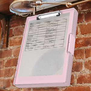 Pink polypropylene clipboard with storage compartment, shown hanging on a brick wall under a light fixture.
