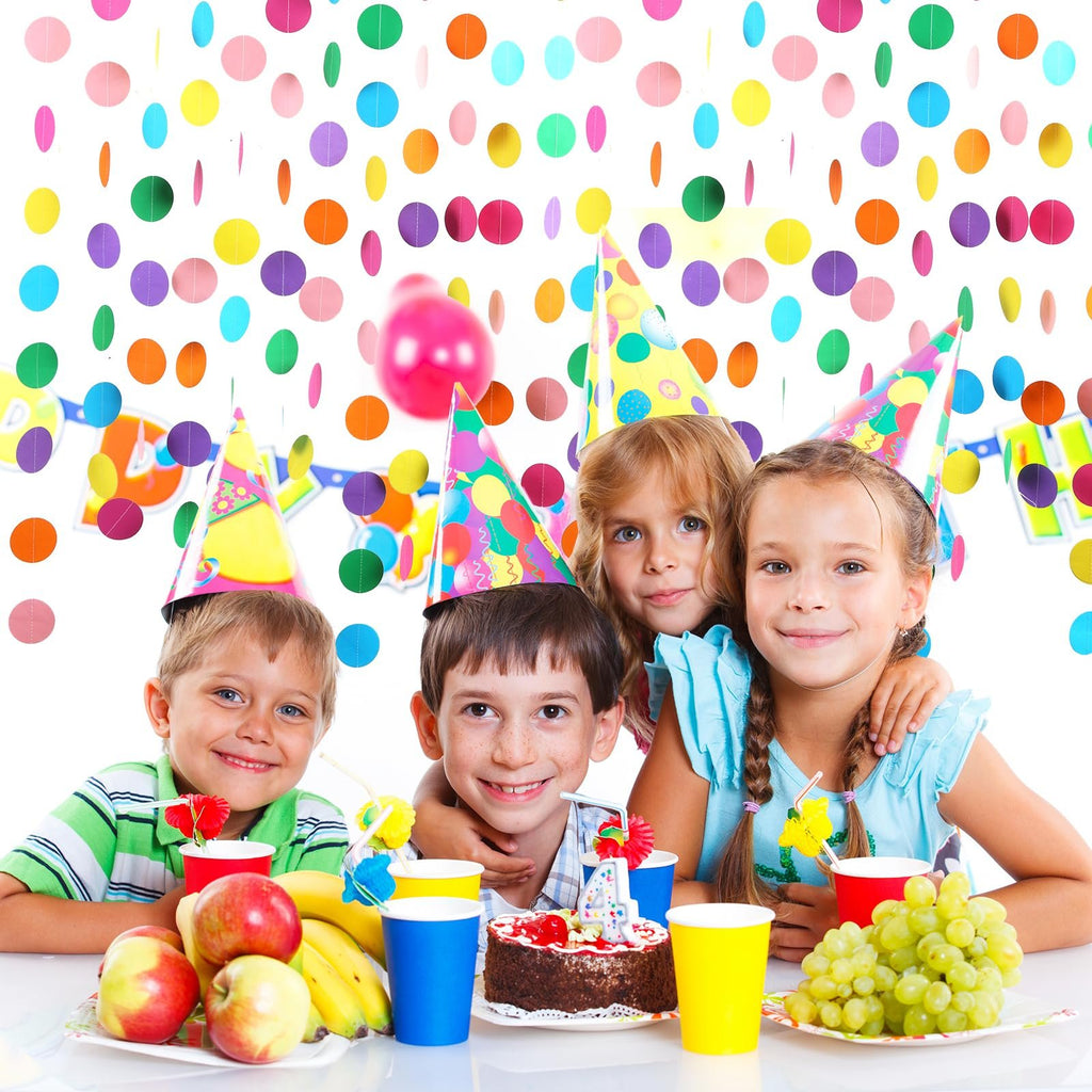 Four smiling children wearing party hats at a table with a cake, fruit, and colorful polka dot decorations