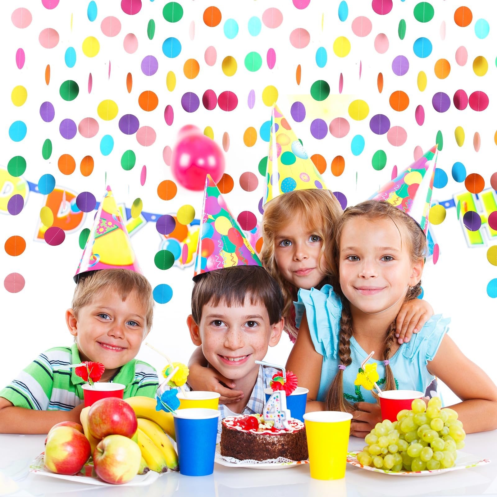Four smiling children wearing party hats at a table with a cake, fruit, and colorful polka dot decorations