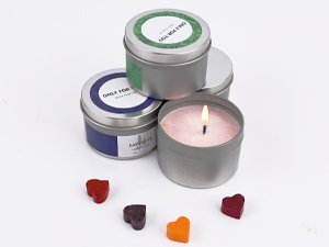 Soy candle making kit contents including four metal tins with labels, one lit candle, and colorful wax heart shapes on a white background.