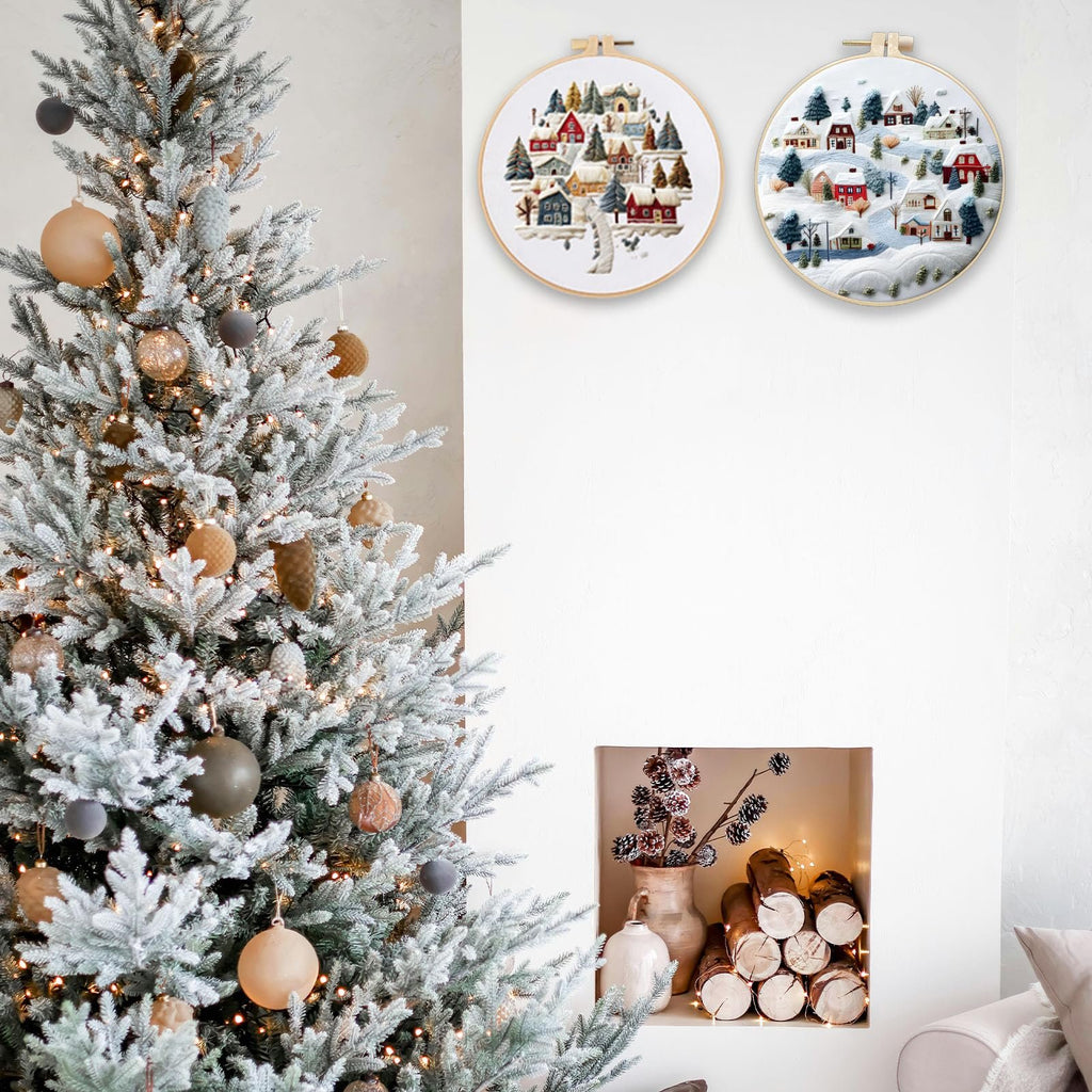 Two Christmas embroidery hoops depicting winter village scenes hang on a wall next to a flocked Christmas tree.