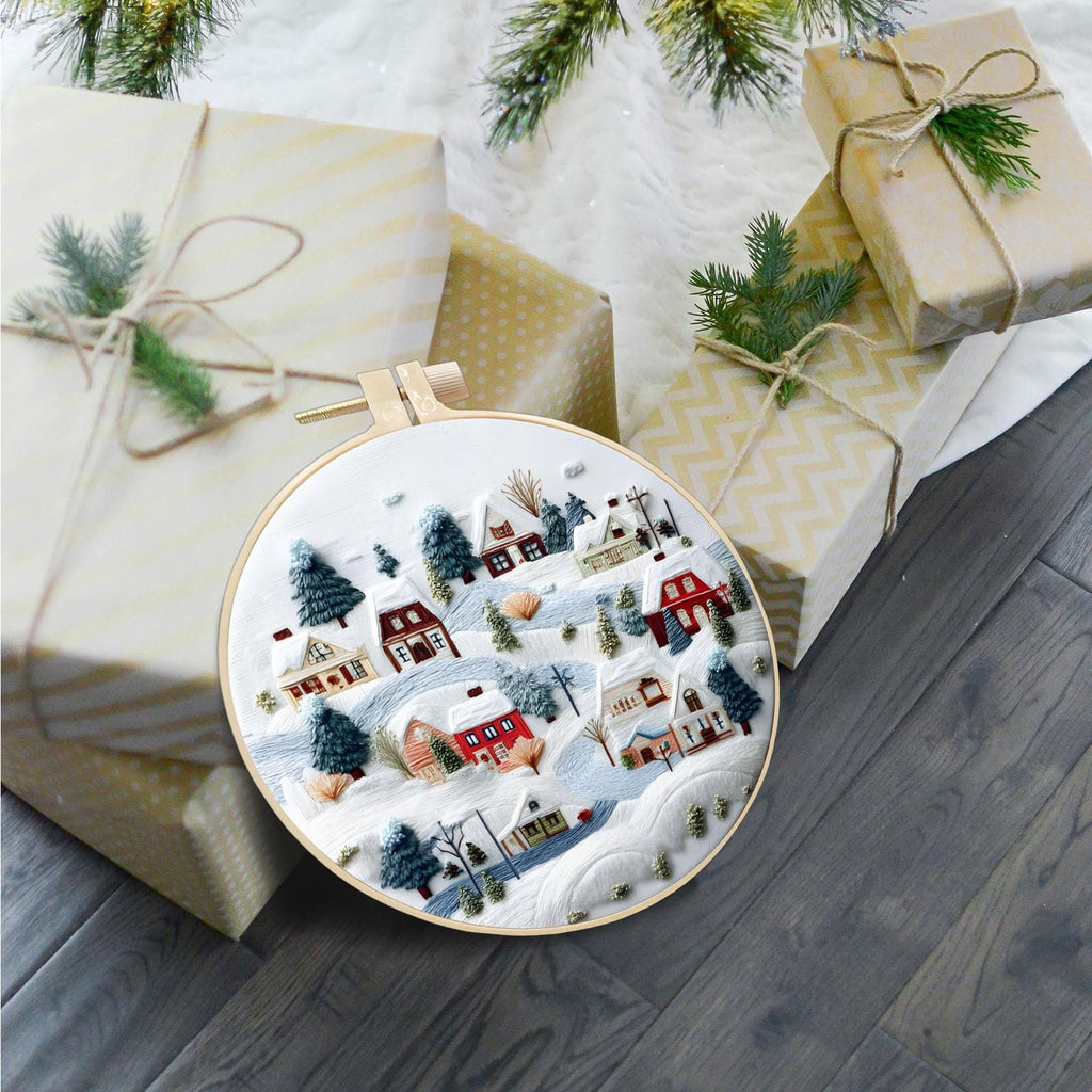 Embroidery hoop with a winter village scene, surrounded by wrapped Christmas gifts and pine branches.