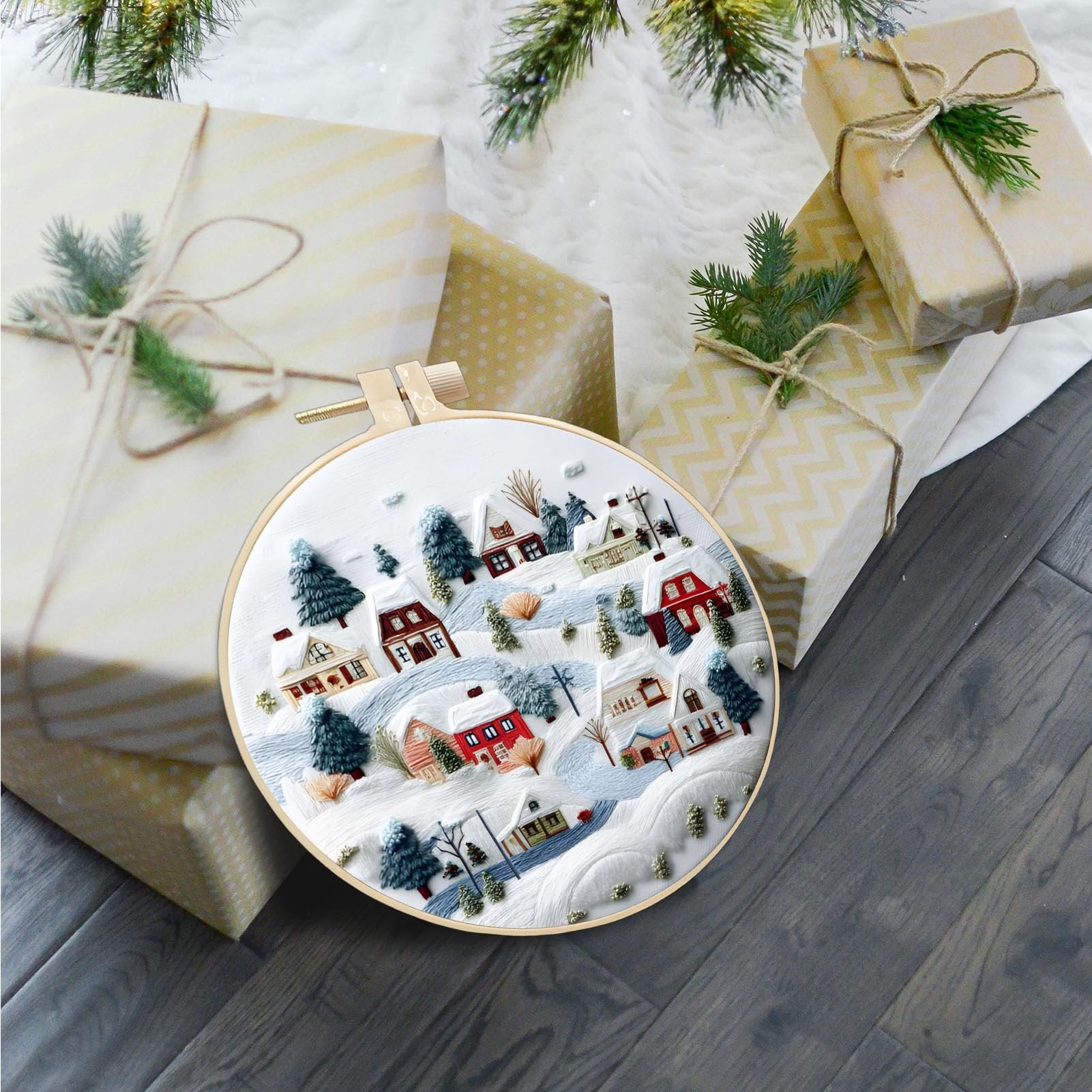 Embroidery hoop with a winter village scene, surrounded by wrapped Christmas gifts and pine branches.