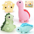 Four crocheted dinosaur toys in various colors, including pink, green, yellow, and blue, with a small box in the background showing illustrations of the toys.