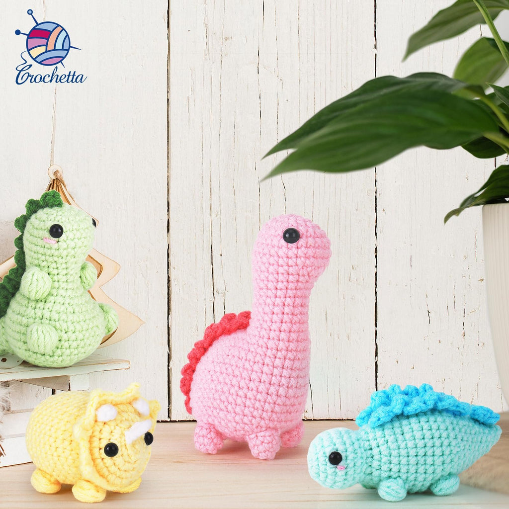 Crocheted dinosaur toys in green, pink, yellow, and blue on a wooden surface with a plant in the background.