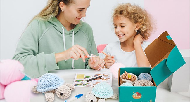 Woman and child crocheting turtle toys from yarn kit, with finished toys and open box of supplies on table.
