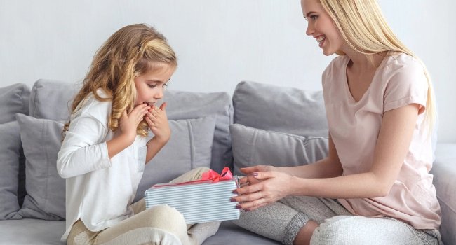 Woman giving a striped gift box with pink ribbon to a surprised young girl on a sofa